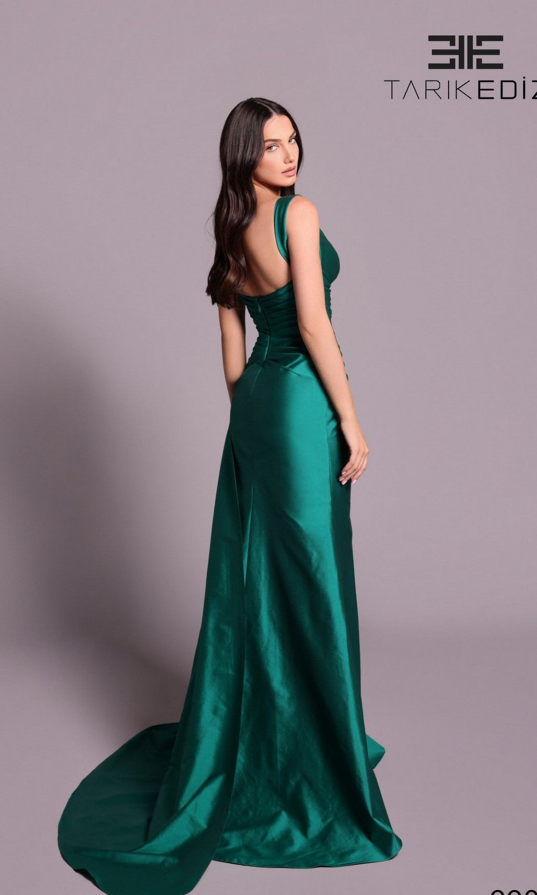 A woman stands with her back turned, glancing over her shoulder in the Long Formal Dress: Tarik Ediz 99035, a floor-length, sleeveless teal satin gown. The